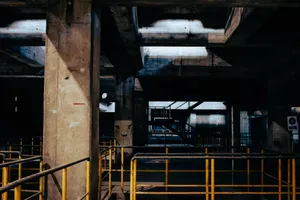 What looks like the inside of an abandoned building, where numerous concrete columns supporting the concrete structure can be seen. There's a hole on the floor between two of the columns, with a metar guardrail around it. At the end, a set of metal stairs is visible. The space is unlit, lending it an apocalyptic atmosphere.