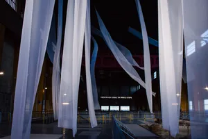 "Prehension" is an art piece by Asad Raza. It features long, white fabric strips hanging off the ceiling that move in the wind, "giv[ing] form to the wind". Behind, there's another piece by Claire Fontaine, consisting of luminous letters that read "When women strike the world stops". The artist wants to defend and reclaim the women's role in history. These two pieces are exhibited on the top floor of the former turbine hall, a large and tall area that also features a giant crane on top. 