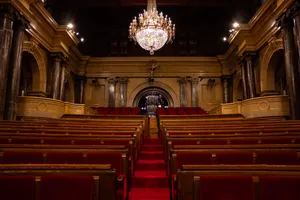 Inside the parliament of Catalonia. It's a surprisingly small room with a narrow central corridor covered with a red carpet and red cushioned seats on both sides of it. Behind the seats, there's a place for visitors and media to sit. At the end of the room, there's a metal clock hanging on the wall. The place is ornamented with ionic style columns and rounded arches. The ceiling, made of wood, has a big ball-like chandelier hanging on.