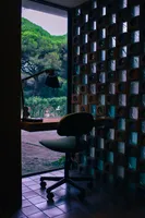 A desk by the window, with a small office chair that faces the outside and a lamp that looks modern but old at the same time, which has a small ashtray mounted on it. On the left side, the wall is made of translucent glass blocks and square bricks with a colored glass circular piece in it, arranged in a checkerboard pattern. Outside, there are some bushes and trees.