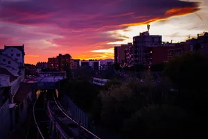 A view of the Mercat Nou metro station, in the lower left corner, alongside the commuter train tracks, under the sunset. A train is leaving the city at the same time as a metro is entering the station, under the sunset.