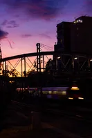 A train speeding beneath a metal pedestrian bridge, under the sunset. The sky has a yellow tone, which fades to purple on top of the photo. The train pictured is a Renfe 447 commuter train, with some graffiti on its side.