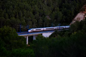 A Velaro E train operated by Renfe speeds up as it exits the city, on top of a bridge in the middle of the forest.