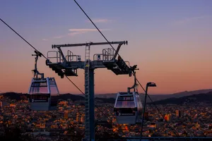 A couple gondolas hanging off the wire, with the sunset behind. The whole city of Barcelona can be seen, as well as the mountains that surround the city.
