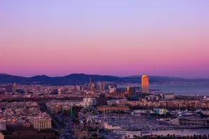 The Barcelona seafront, under the sunset. There's a big Ferris wheel next to the Olympic port, by the seashore. There are also a couple tall skyscrapers by the sea. The mountains can be seen on the back.