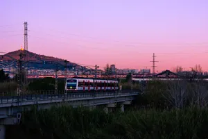 An FGC class 213 narrow-gauge train on its way by Sant Boi, passing by a concrete bridge across the Llobregat river. On the background, a Frecciarossa high speed train speeds up on an elevated viaduct.