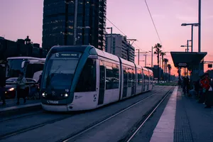 A tramway stationed at a tram stop in front of a tall and black skyscraper. The sky behind is bright orange. The rest of the image has desaturated colors.