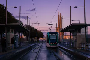 A green Barcelona tramway leaving its stop. The ground underneath is still wet, as it had just rained. At the background, the three chimneys of Sant Adrià can be seen.