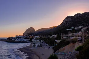 A tranquil town surrounded by mountains by the Catalonian coast, by sunset. There's a small and quiet beach by the sea. A train runs through the town on elevated tracks, on the right of the shot.