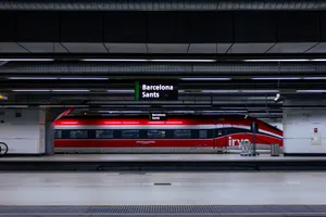A striking red ETR 1000 "Frecciarossa" high speed train, stationed at Barcelona Sants train station. The picture, captured from several platforms away, shows the side of the train, which highlights the aerodynamic design the train has.