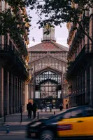 The Born market, now turned into a museum. It was the first big building constructed using an iron structure in Barcelona. Inspired in the late 19th century French markets, it offered a large commercial space using only little material. Below the surface, the remnants of the old Born neighbourhood, destroyed during the early 18th century by the Bourbonic army, were found after the market came to a close. The side entrance can be seen through a narrow street, with two art nouveau style buildings on each side. A black-and-yellow taxi is speeding through the main road perpendicular to the street.